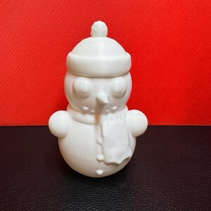 White 3D-printed snowman LED tealight holder shown against a bold red background, highlighting its intricate winter-themed details like the scarf, buttons, and hat.
