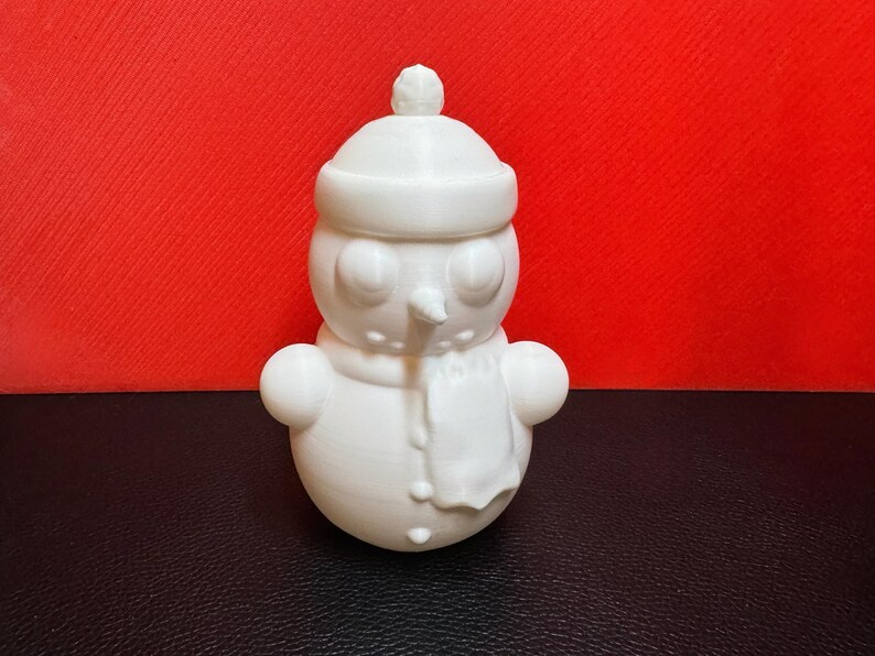 White 3D-printed snowman LED tealight holder shown against a bold red background, highlighting its intricate winter-themed details like the scarf, buttons, and hat.