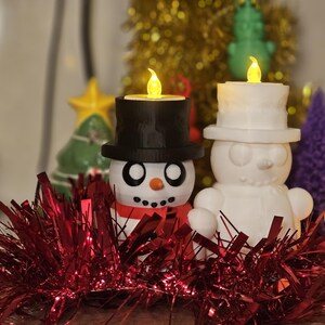 Adorable 3D-printed snowman tealight holders in white and multicolour, featuring festive designs with hats, scarves, and LED tealights. Perfect as Christmas decorations or gifts, displayed on a table with holiday-themed backdrops.