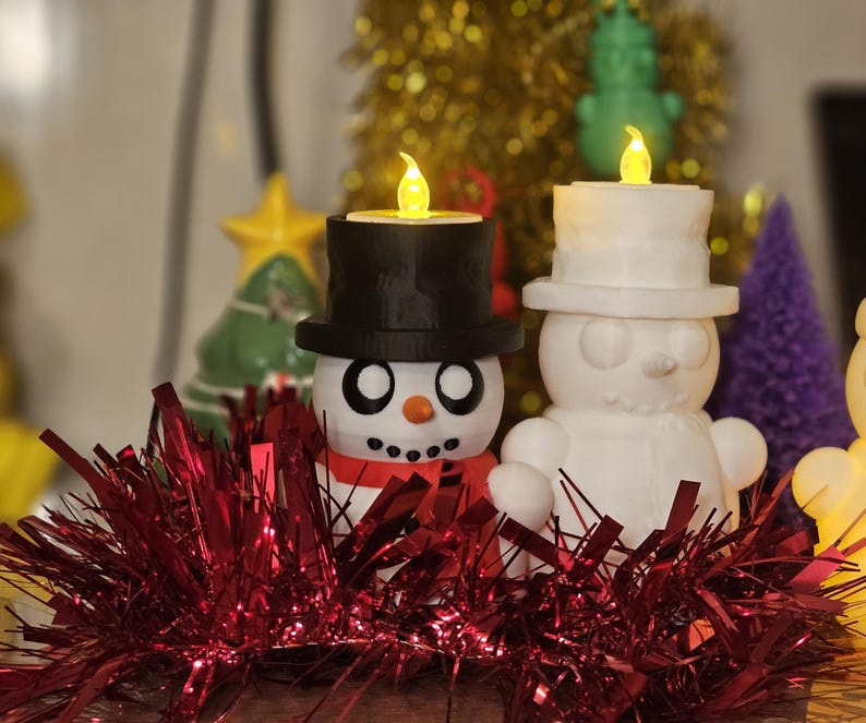 Adorable 3D-printed snowman tealight holders in white and multicolour, featuring festive designs with hats, scarves, and LED tealights. Perfect as Christmas decorations or gifts, displayed on a table with holiday-themed backdrops.