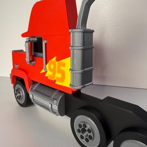 May include: A red and yellow toy semi-truck with the number 95 on the side. The truck has a black trailer and silver wheels. The exhaust pipe is gray. The toy is a replica of a cartoon character.