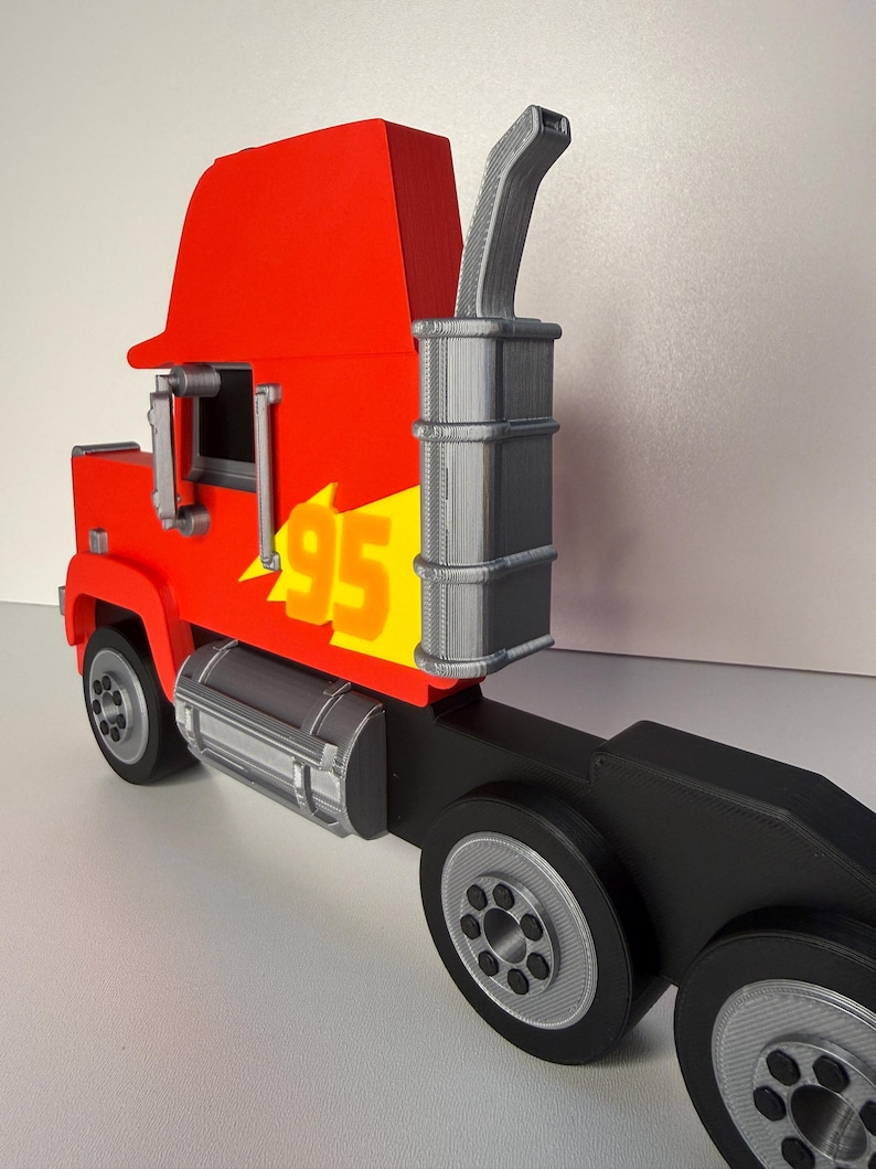 May include: A red and yellow toy semi-truck with the number 95 on the side. The truck has a black trailer and silver wheels. The exhaust pipe is gray. The toy is a replica of a cartoon character.