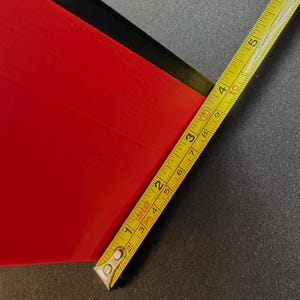 May include: A red rectangular object with a black edge is measured with a yellow tape measure. The tape measure shows measurements in inches and feet. The object appears to be approximately 3 inches wide.