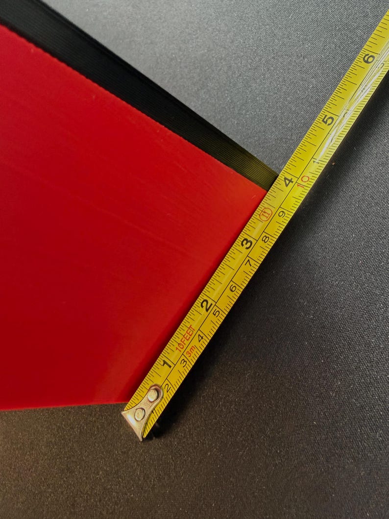 May include: A red rectangular object with a black edge is measured with a yellow tape measure. The tape measure shows measurements in inches and feet. The object appears to be approximately 3 inches wide.