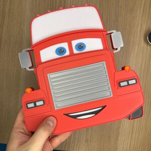 May include: Red cartoon truck-shaped item with a white top, blue eyes, and a smiling mouth. The truck has a silver grill and side mirrors. The item is held by a hand.
