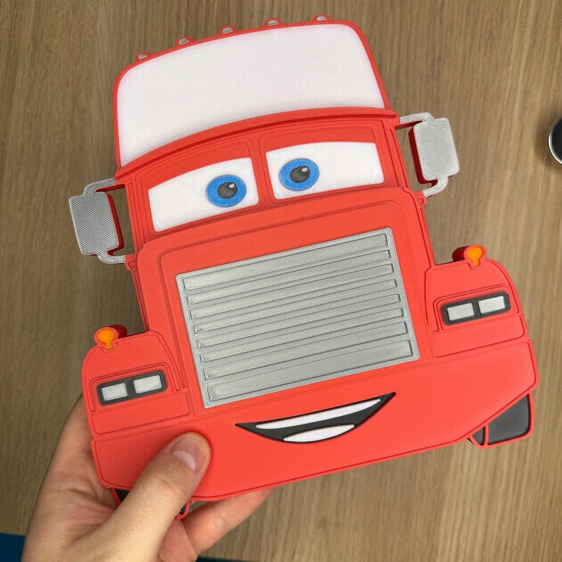 May include: Red cartoon truck-shaped item with a white top, blue eyes, and a smiling mouth. The truck has a silver grill and side mirrors. The item is held by a hand.
