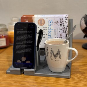 bedside organiser with a book stand, phone holder, pen storage, and a cup holder. Shown in grey with a book, hot drink, and phone displaying a sleep tracking app. Ideal for nightstands, keeping essentials tidy and within reach