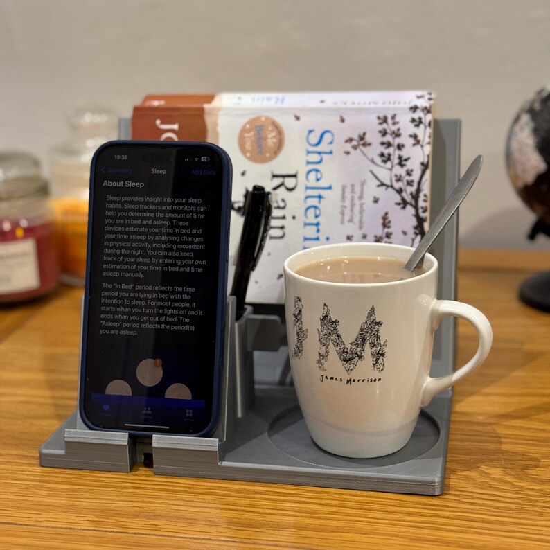 bedside organiser with a book stand, phone holder, pen storage, and a cup holder. Shown in grey with a book, hot drink, and phone displaying a sleep tracking app. Ideal for nightstands, keeping essentials tidy and within reach