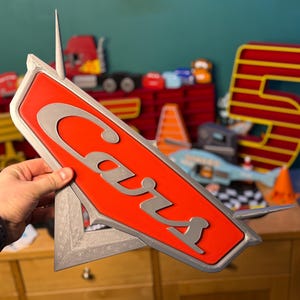 May include: A red and silver Cars logo sign. The word "Cars" is in silver, stylized lettering on a red background. The sign has a silver border and a pointed top. Other toy cars and a number 5 are visible in the background.