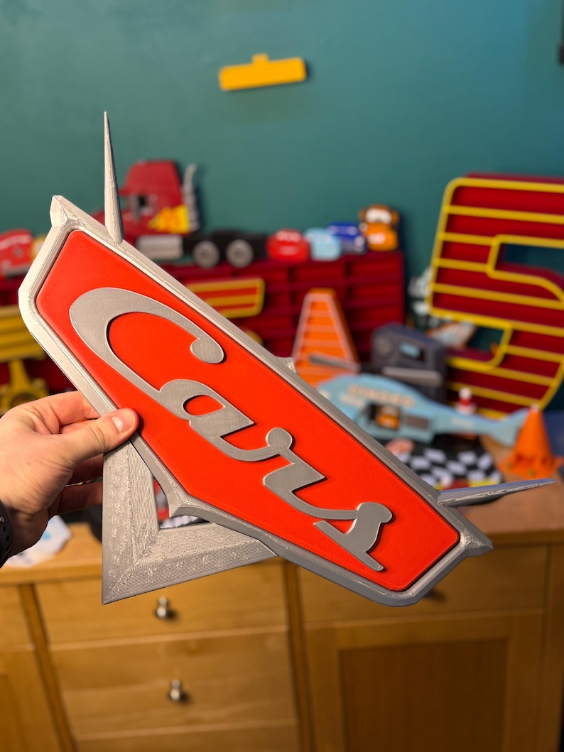 May include: A red and silver Cars logo sign. The word "Cars" is in silver, stylized lettering on a red background. The sign has a silver border and a pointed top. Other toy cars and a number 5 are visible in the background.