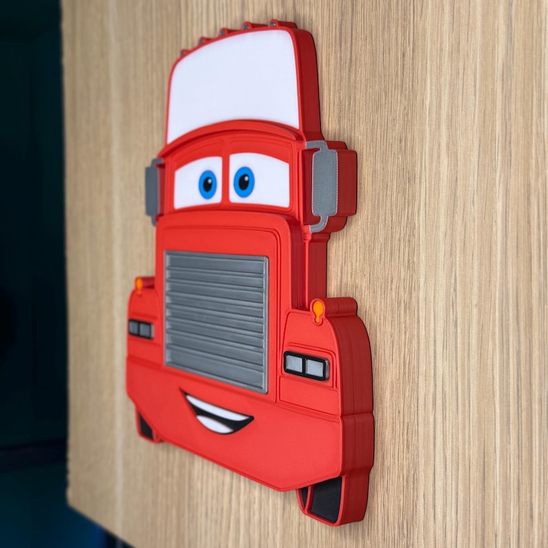 May include: A red truck-shaped wall decoration featuring a white roof, a grey grille, and blue eyes. The truck has a smiling mouth, orange headlights, and black accents. This three-dimensional object is designed with a cartoon-like appearance.