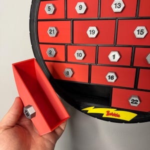 May include: Red and black advent calendar with silver numbered compartments. A red drawer is open, held by a hand, revealing a silver number 12. The word "Bobbie" is printed in yellow.