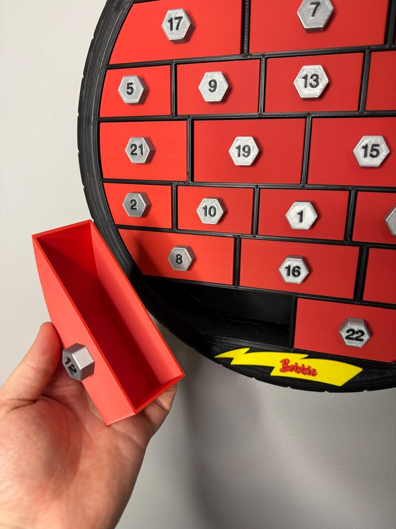 May include: Red and black advent calendar with silver numbered compartments. A red drawer is open, held by a hand, revealing a silver number 12. The word "Bobbie" is printed in yellow.