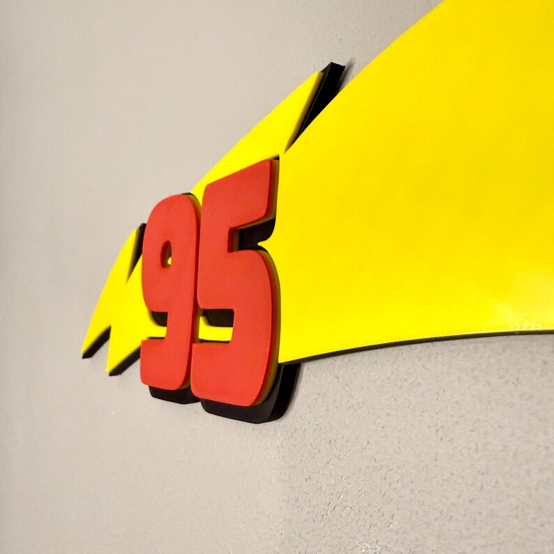 May include: A wall decoration featuring the number "95" in red, with a black outline, set against a yellow lightning bolt shape. The design is three-dimensional and mounted on a light gray wall.