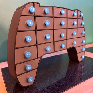 May include: A brown, game controller-shaped advent calendar with 24 small drawers. Each drawer has a light blue knob with a number printed on it. The advent calendar is made of wood and sits on a dark surface.