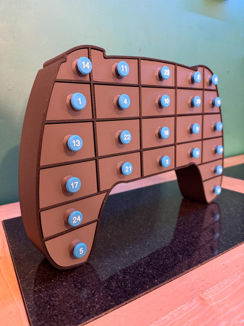 May include: A brown, game controller-shaped advent calendar with 24 small drawers. Each drawer has a light blue knob with a number printed on it. The advent calendar is made of wood and sits on a dark surface.