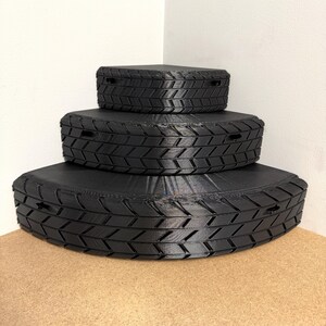 May include: Three black, tire-shaped storage boxes stacked on top of each other. The boxes have a textured tread pattern and small rectangular openings. The boxes are on a light brown surface, with a white wall in the background.