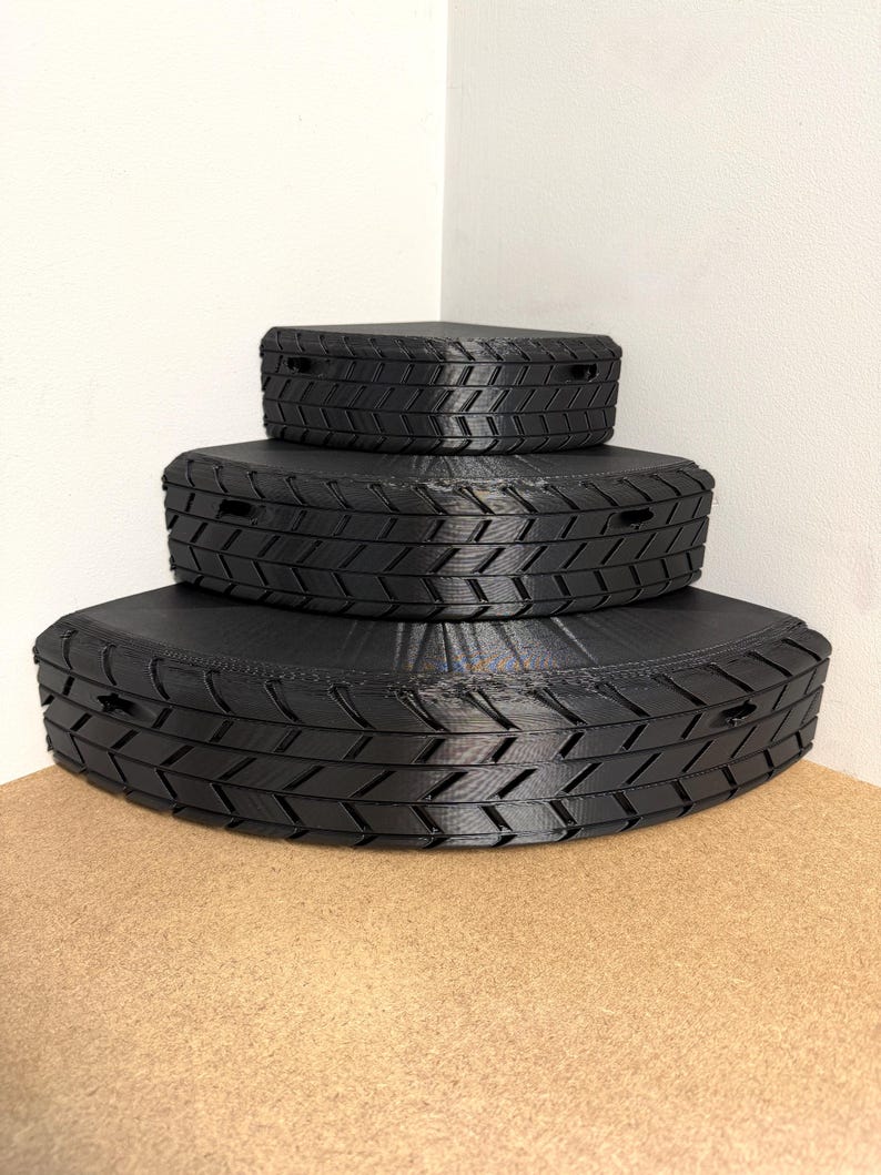 May include: Three black, tire-shaped storage boxes stacked on top of each other. The boxes have a textured tread pattern and small rectangular openings. The boxes are on a light brown surface, with a white wall in the background.