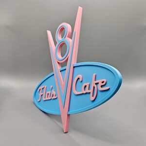 May include: A retro-style sign for "Fla's Cafe" in pink and blue. The sign features a stylized "V8" design above an oval with the cafe's name. The color scheme is reminiscent of classic diner aesthetics.