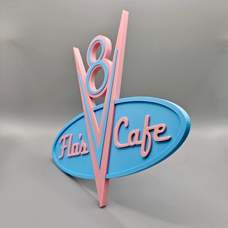 May include: A retro-style sign for "Fla's Cafe" in pink and blue. The sign features a stylized "V8" design above an oval with the cafe's name. The color scheme is reminiscent of classic diner aesthetics.