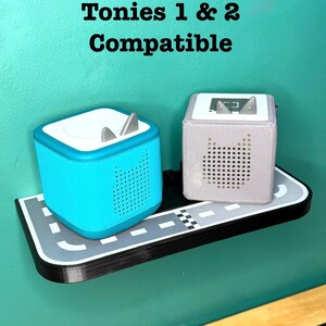 May include: Two Tonies audio players, one turquoise and one light purple, sit on a shelf designed as a road. The text "Tonies 1 & 2 Compatible" is above the players. The turquoise player has silver ears.