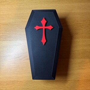 Top down view of Black 3D-printed reptile hide shaped like a gothic coffin with a raised red cross on top. 9 inches in length. Features an arched entrance on one side, designed for snakes, geckos, or other small reptiles. Placed on a wooden surface.