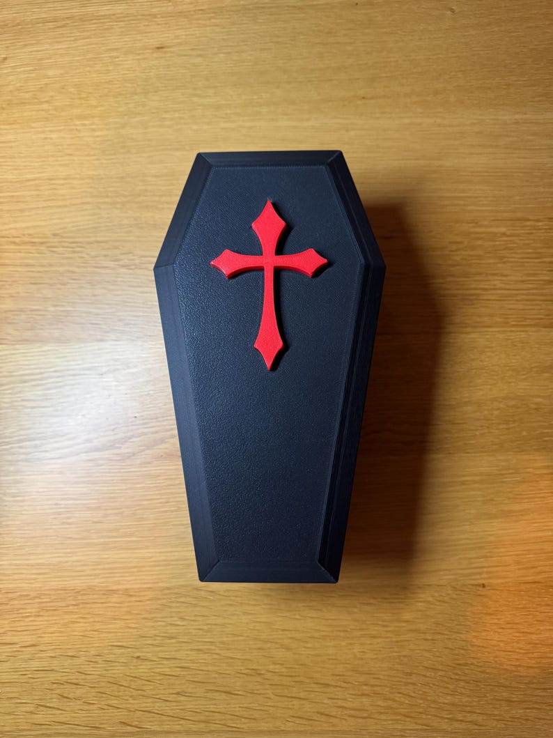 Top down view of Black 3D-printed reptile hide shaped like a gothic coffin with a raised red cross on top. 9 inches in length. Features an arched entrance on one side, designed for snakes, geckos, or other small reptiles. Placed on a wooden surface.