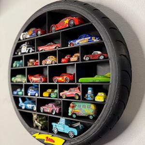 May include: Black, circular display shelf made from a tire, filled with various toy cars. The cars are in different colors and styles, including race cars, trucks, and vans. The shelf is mounted on a white wall.