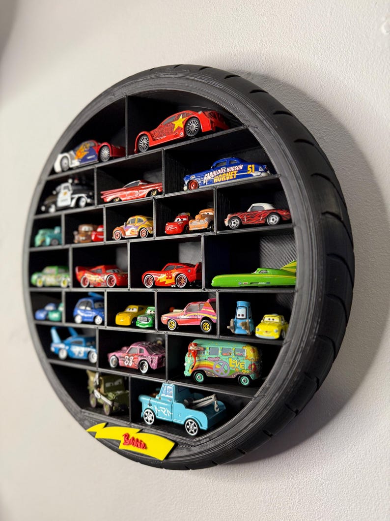 May include: Black, circular display shelf made from a tire, filled with various toy cars. The cars are in different colors and styles, including race cars, trucks, and vans. The shelf is mounted on a white wall.