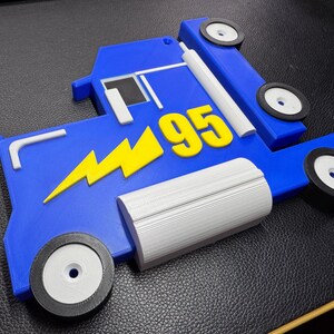 A 3D-printed blue truck model inspired by Mack from Cars, featuring a yellow lightning bolt and the number 95. The parts are laid flat, showing two wheels, grey detailing, and black tires, all on a leather surface.