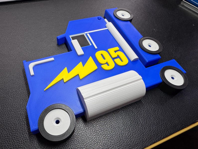A 3D-printed blue truck model inspired by Mack from Cars, featuring a yellow lightning bolt and the number 95. The parts are laid flat, showing two wheels, grey detailing, and black tires, all on a leather surface.
