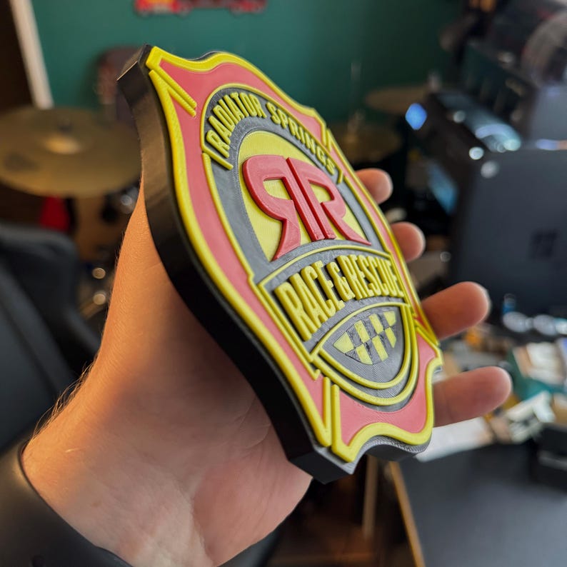 May include: A multi-layered, shield-shaped emblem with the text "RADIATOR SPRINGS RACE & RESCUE". The emblem features red, yellow, gray, and black colors. The emblem is held in a hand.