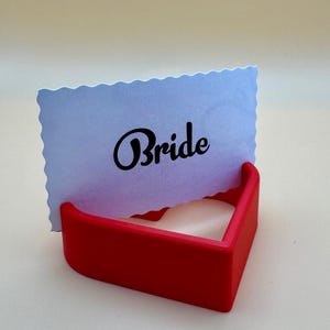 Red 3D-printed heart-shaped place card holder displaying a “Bride” card. Perfect for weddings, bridal showers, or receptions, this durable and elegant design adds charm to table settings and complements romantic themes for special events.