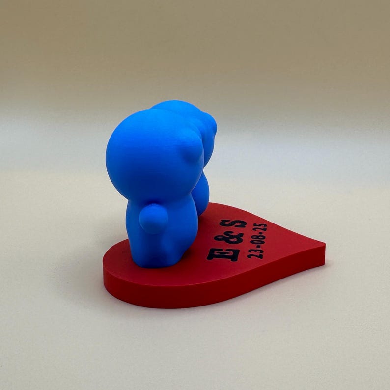 Cute 3D-printed cake topper featuring two blue figurines side by side on a red heart base engraved with initials and a date. Ideal for weddings, anniversaries, or events, this cute eco-friendly PLA decoration adds charm to any special occasion.