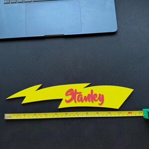 May include: A yellow lightning bolt-shaped sign with the name "Stanley" in red lettering. The sign is approximately 13 inches long, as measured by a yellow tape measure. The background is black.