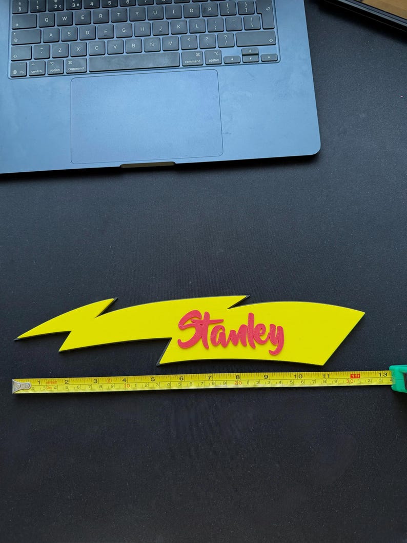 May include: A yellow lightning bolt-shaped sign with the name "Stanley" in red lettering. The sign is approximately 13 inches long, as measured by a yellow tape measure. The background is black.