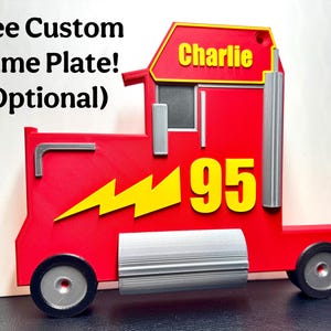 A 3D-printed red truck display inspired by Mack from Cars, featuring a bold yellow “95” and a lightning bolt. Silver details add realism, and a custom nameplate option is glued neatly on, fitting the profile of truck, reading “Charlie.”