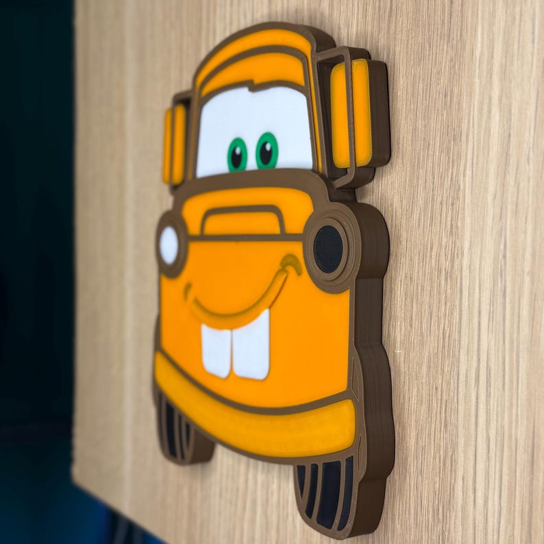 May include: A 3D wall art piece featuring the character Mater from the animated movie Cars. The artwork is primarily orange with brown and white accents, including Mater's signature buck teeth and smiling face. The piece is mounted on a wooden surface.