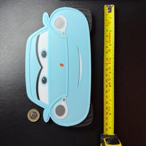 May include: A light blue car-shaped object with black tires and white and black details. A yellow tape measure shows the object is approximately 9 inches long. A coin is next to the car.