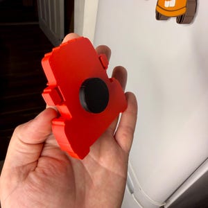 May include: A red magnet shaped like a car, held in a hand. The magnet has a black circular magnet attached to the back. Other car-shaped magnets are visible in the background.