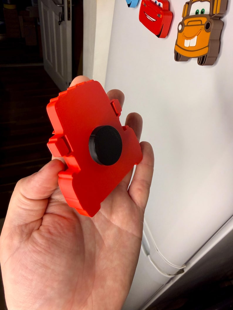 May include: A red magnet shaped like a car, held in a hand. The magnet has a black circular magnet attached to the back. Other car-shaped magnets are visible in the background.
