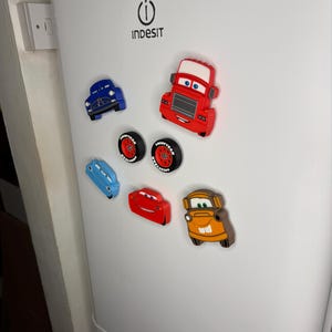 May include: A collection of colorful car-shaped magnets, including blue, red, and orange vehicles, and two tire magnets. The magnets are attached to a white refrigerator door. The magnets are designed to look like characters from the movie Cars.