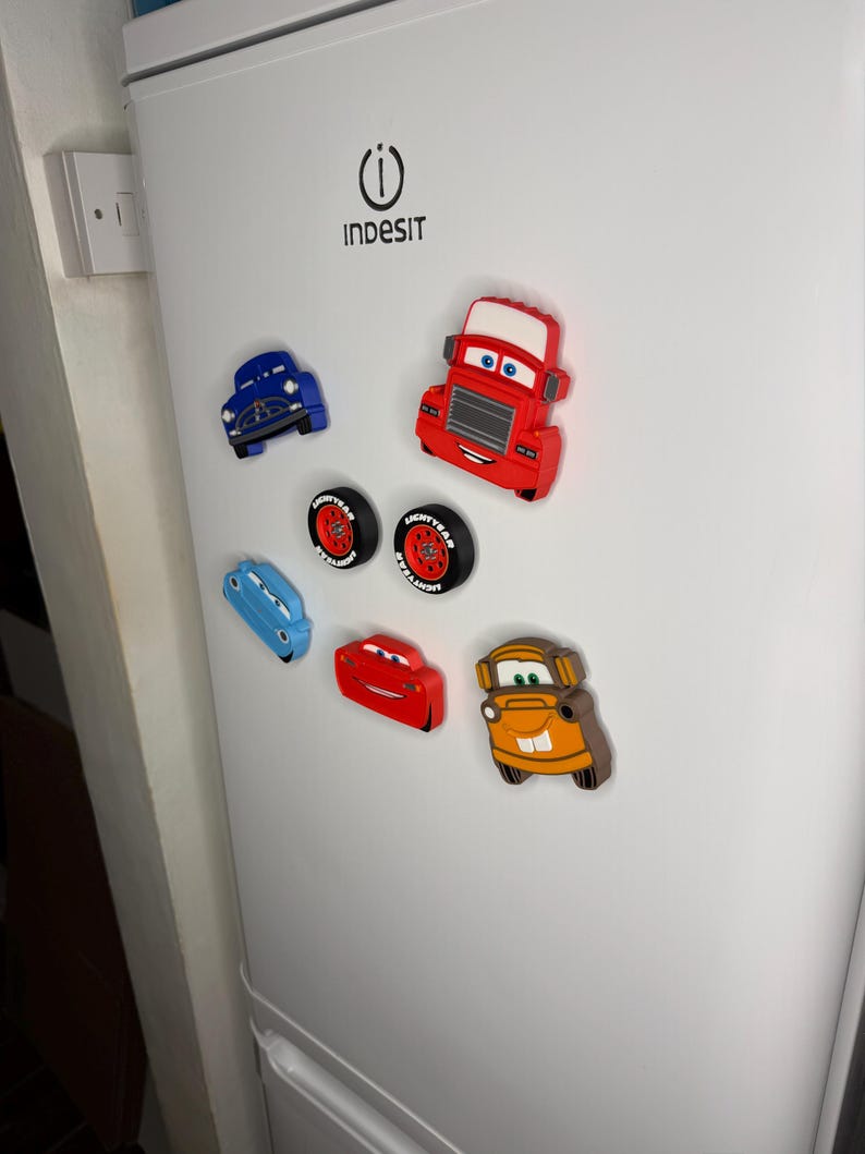 May include: A collection of colorful car-shaped magnets, including blue, red, and orange vehicles, and two tire magnets. The magnets are attached to a white refrigerator door. The magnets are designed to look like characters from the movie Cars.