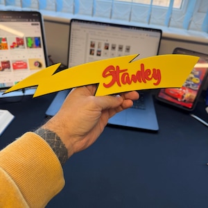 May include: A yellow lightning bolt-shaped sign with the name "Stanley" in red, held by a person. The sign has a black outline and is likely made of wood or a similar material. The background includes a laptop, tablet, and other office items.
