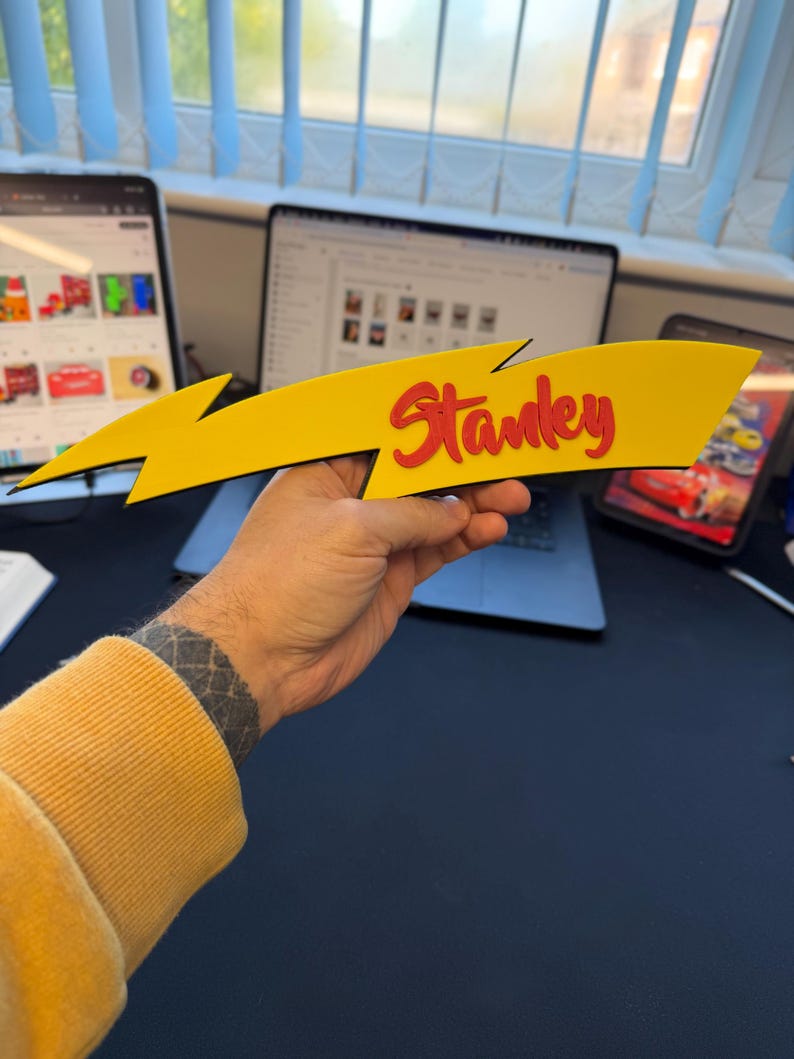 May include: A yellow lightning bolt-shaped sign with the name "Stanley" in red, held by a person. The sign has a black outline and is likely made of wood or a similar material. The background includes a laptop, tablet, and other office items.