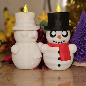 Adorable 3D-printed snowman tealight holders in white and multicolour, featuring festive designs with hats, scarves, and LED tealights. Perfect as Christmas decorations or gifts, displayed on a table with holiday-themed backdrops.