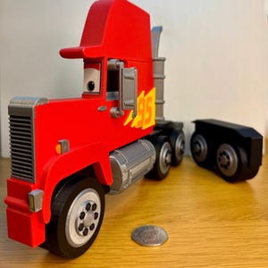 May include: A red toy semi-truck with a black trailer. The truck features a red cab with a visor, a silver grill, and the number "95" in yellow with a flame design. The wheels are black with silver rims. A coin is next to the truck.