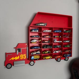 May include: A red toy truck with a number 95 on the side, with a red and white lightning bolt design. The truck has a shelf on the back that holds 18 small toy cars. The shelf is divided into 18 compartments, each holding a different toy car.