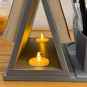 close-up of the book stand section with two LED tea lights placed inside, creating a warm glow. The silver 3D-printed structure holds an open book, showing its dual function as a reading light and decorative nightstand piece
