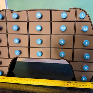 May include: A brown, multi-compartment advent calendar with blue knobs, numbered 1-24. The calendar is shaped like a rounded rectangle. A yellow tape measure shows a length of approximately 16 inches.